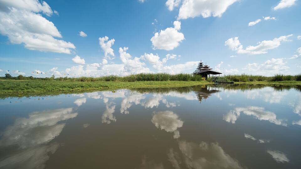 A serene pond with a pagoda reflecting in the water, surrounded by lush greenery and a clear blue sky with scattered clouds.