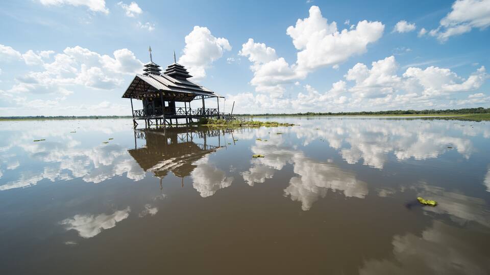 A traditional stilt house with a reflection in calm water, surrounded by a clear sky and distant greenery.