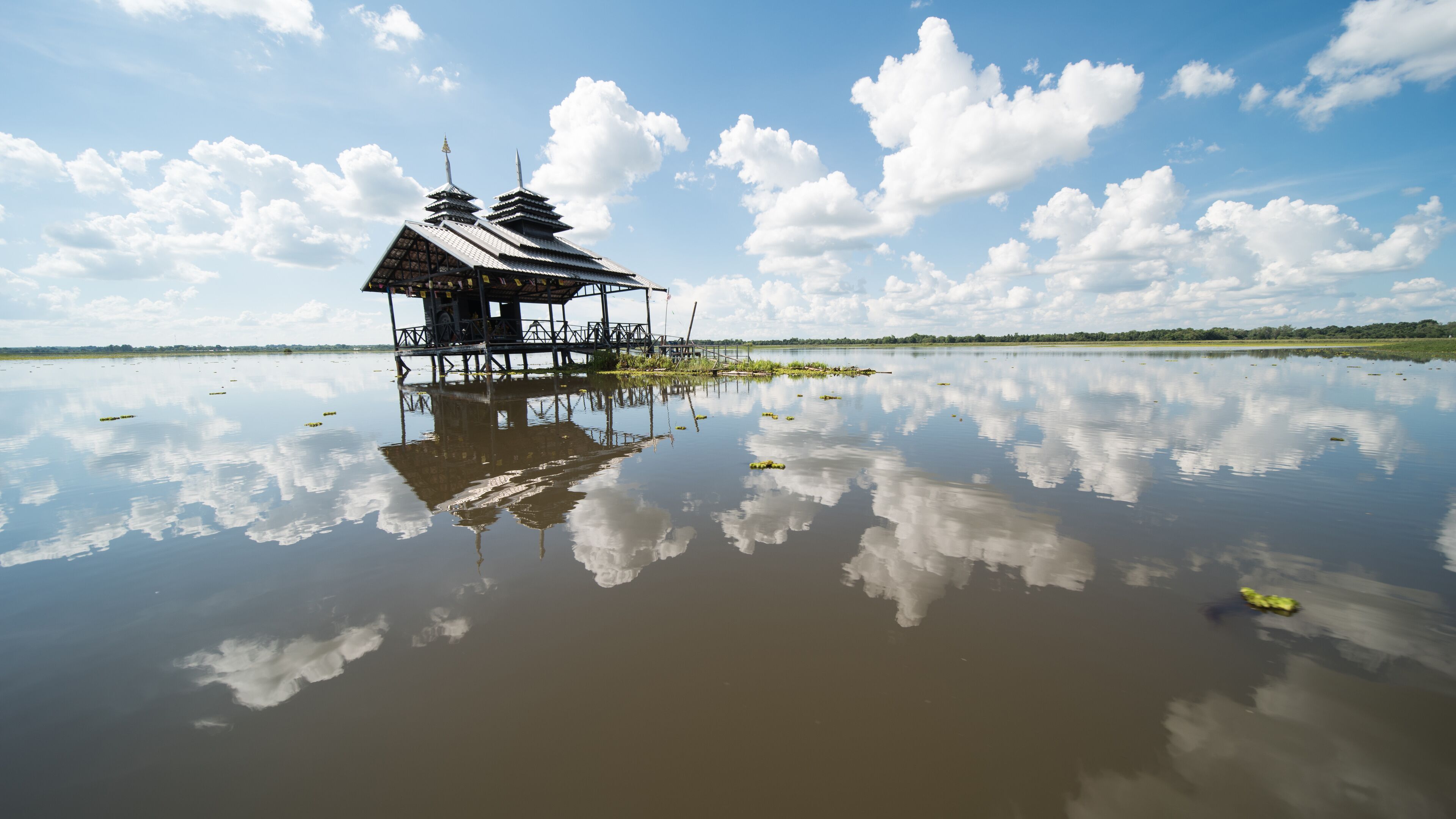 A traditional stilt house with a reflection in calm water, surrounded by a clear sky and distant greenery.