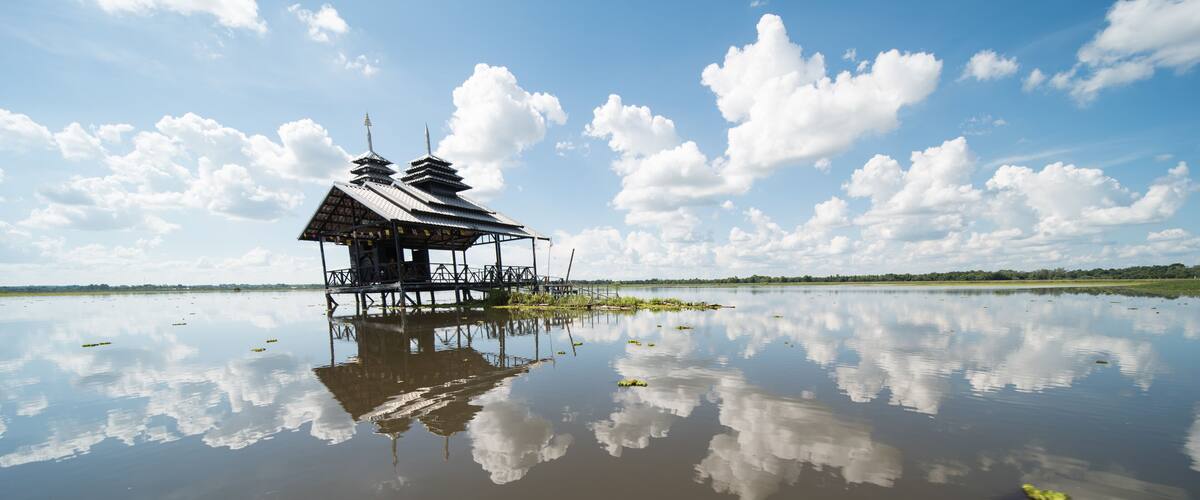 A traditional stilt house with a reflection in calm water, surrounded by a clear sky and distant greenery.