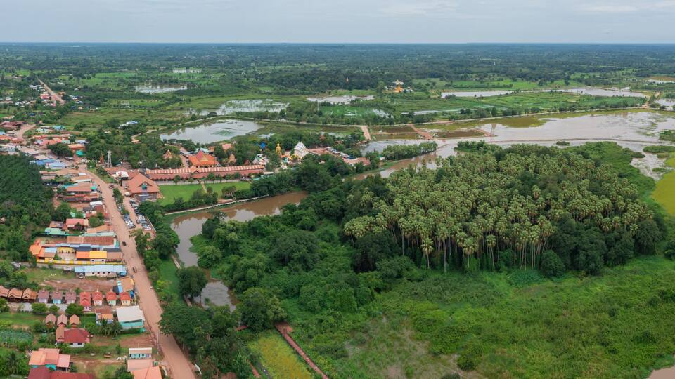 An aerial view of a rural area with a river, dense forest, and a village with colorful houses.