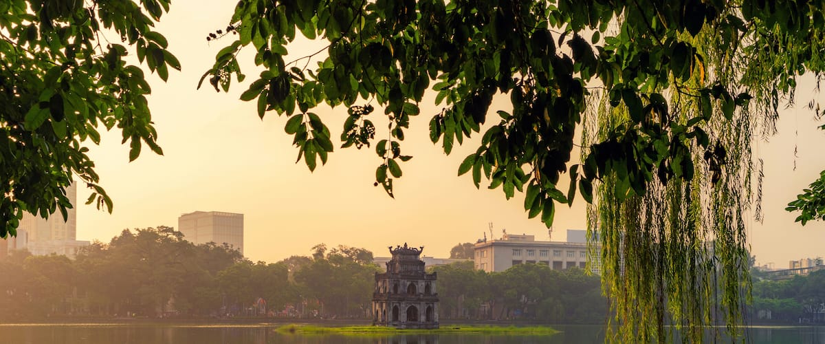 A serene lake with a traditional pagoda in the center, surrounded by lush greenery and modern buildings in the background.