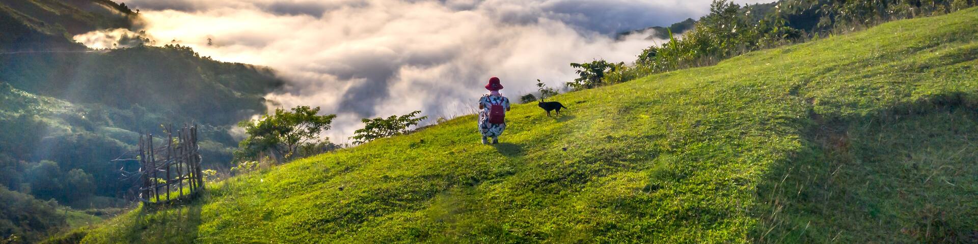 A person walking on a grassy hillside with a mountain range in the background and clouds below.