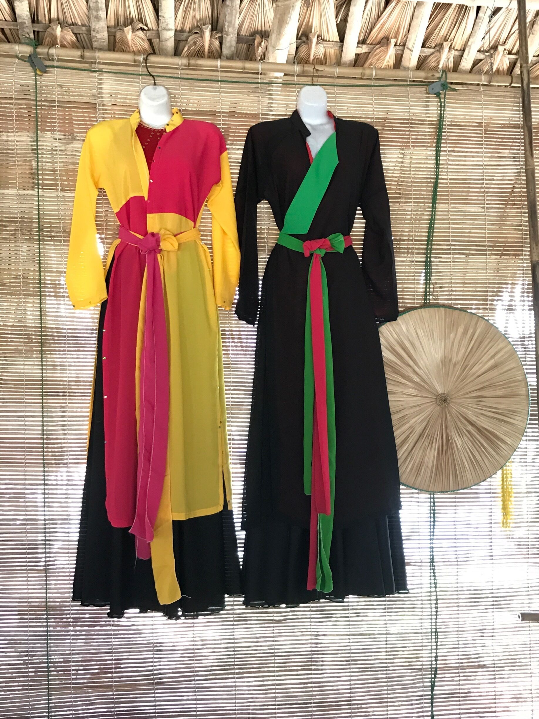Two mannequins dressed in colorful dresses with long sleeves and belts, against a bamboo backdrop.