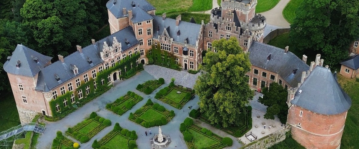 Vista aérea de un castillo grande e histórico con un jardín formal, rodeado de campos verdes exuberantes y árboles.