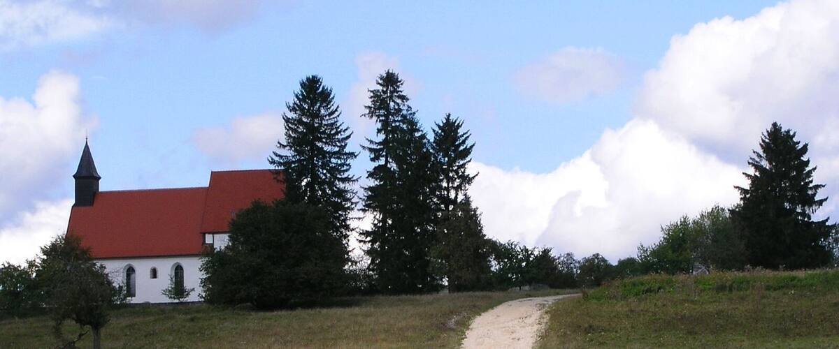 Eine Kirche mit rotem Dach und weißen Wänden, umgeben von Bäumen und einem Feldweg.