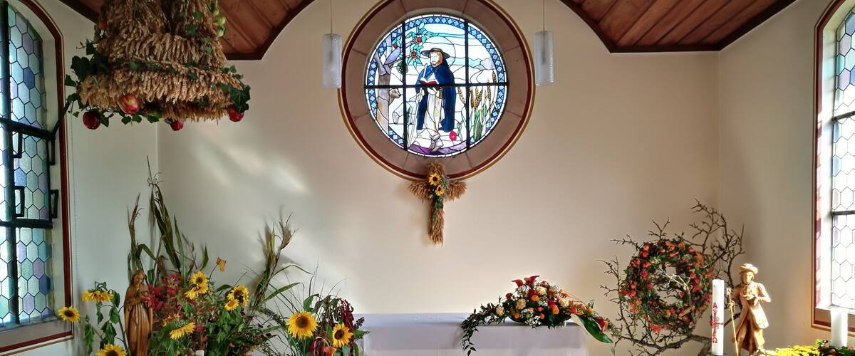 A church interior with a wooden ceiling, a stained glass window depicting a religious figure, and various floral arrangements.