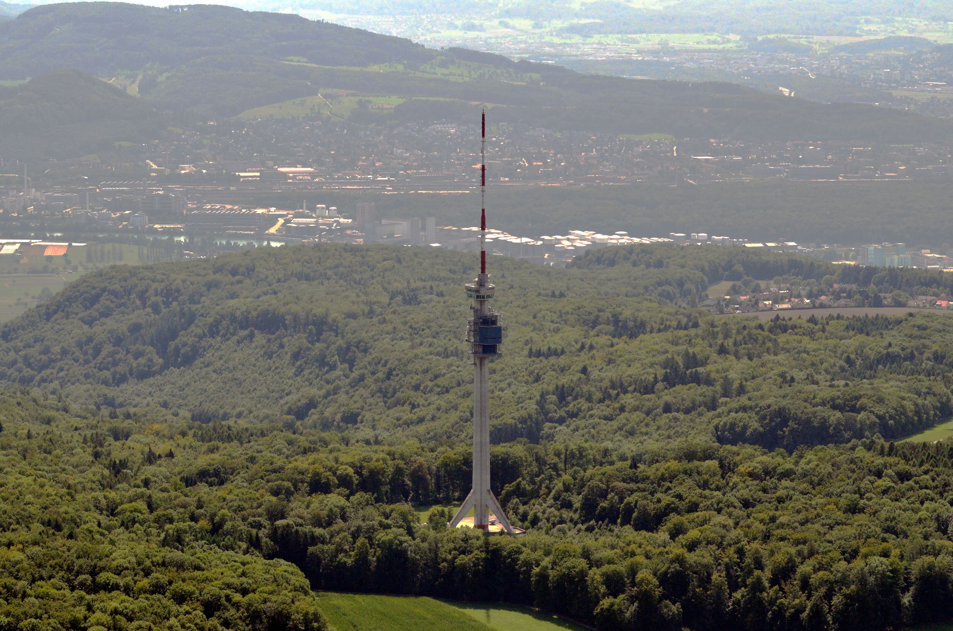 A tall communication tower surrounded by dense forest and a distant cityscape.