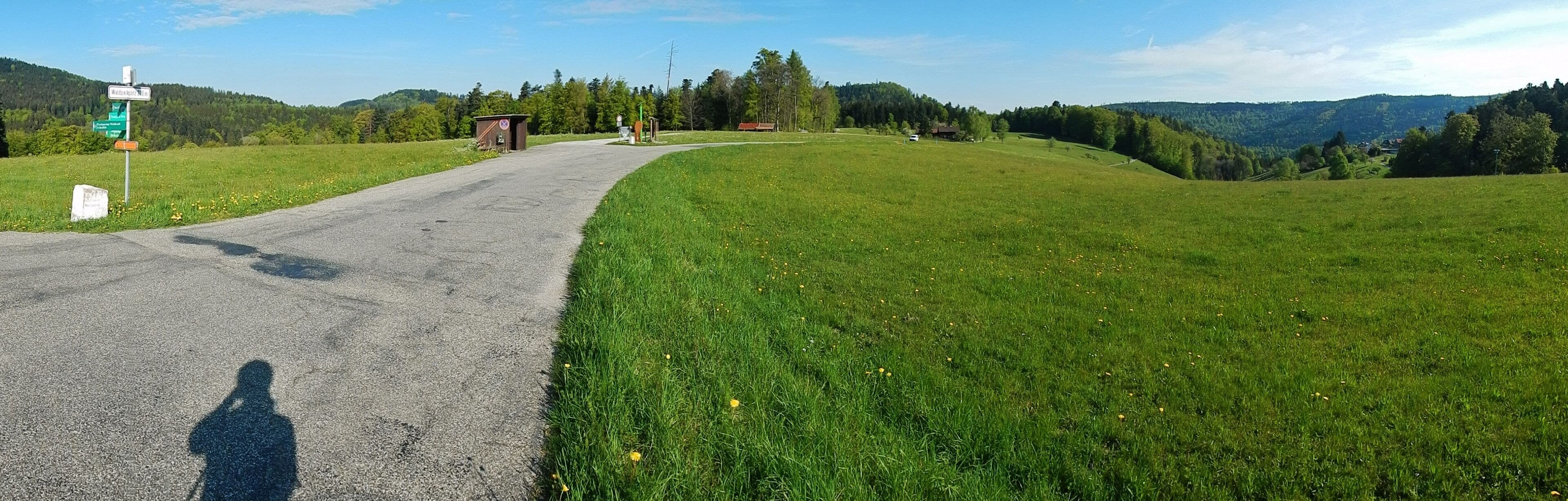 A rural road curving through a green field with a small building and a signpost in the distance.