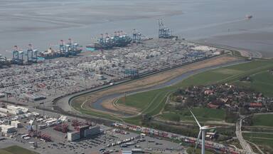An industrial port with shipping containers, cranes, and a wind turbine.