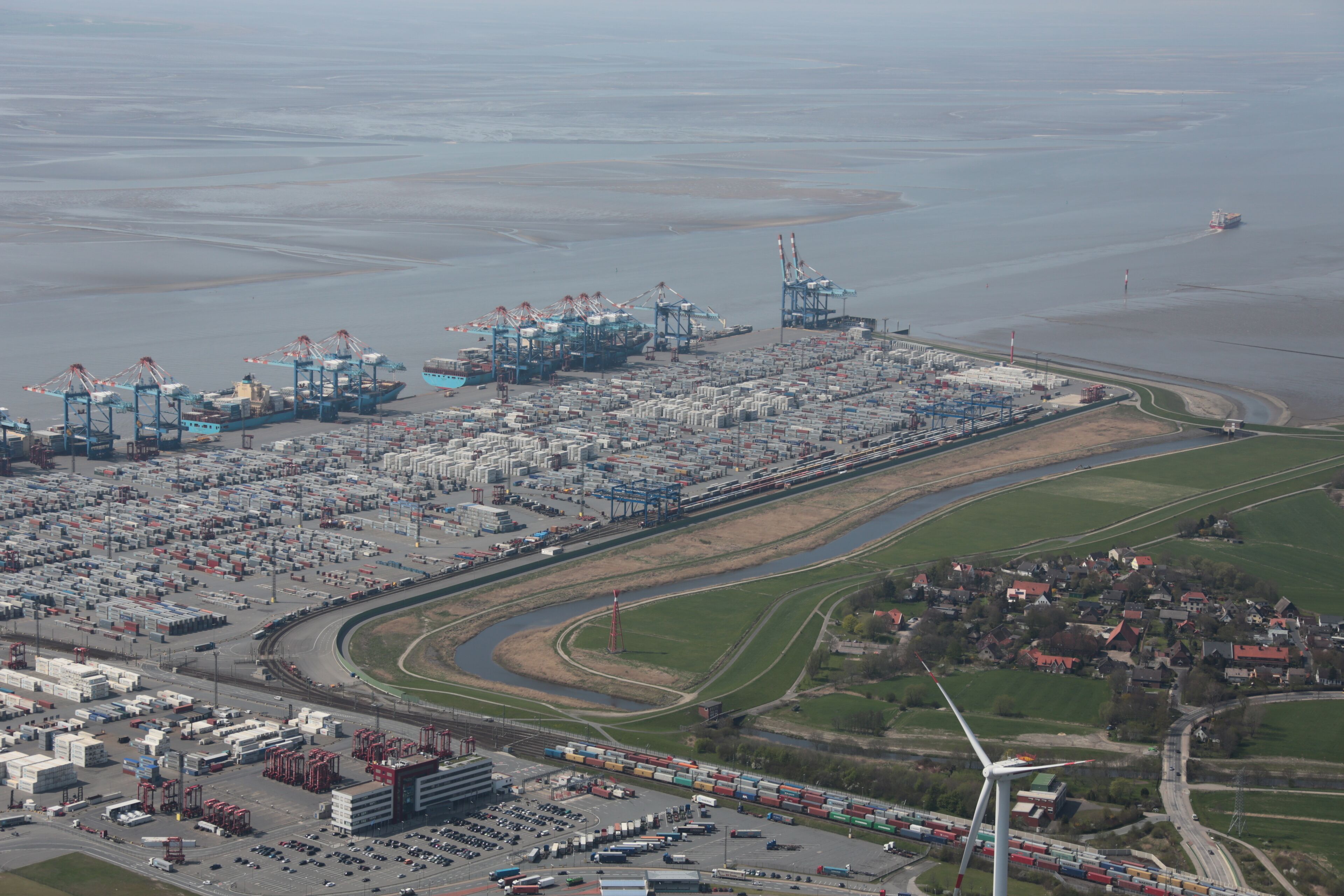 An industrial port with shipping containers, cranes, and a wind turbine.