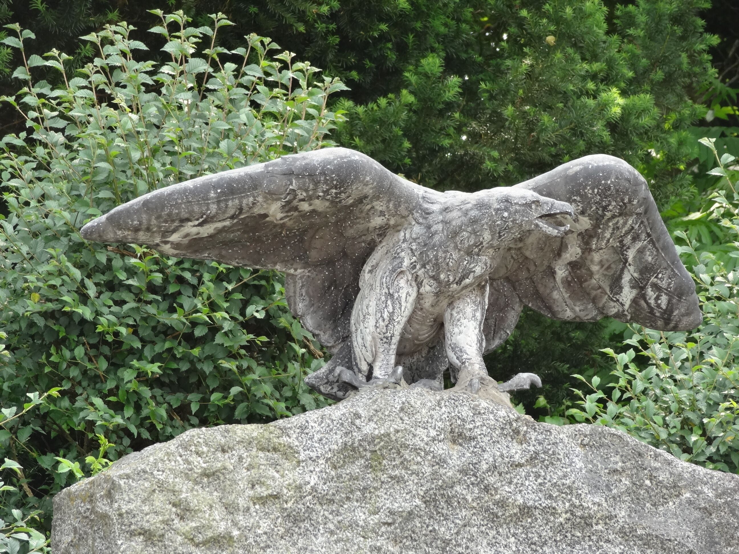 A stone eagle statue on a rock with green foliage in the background.
