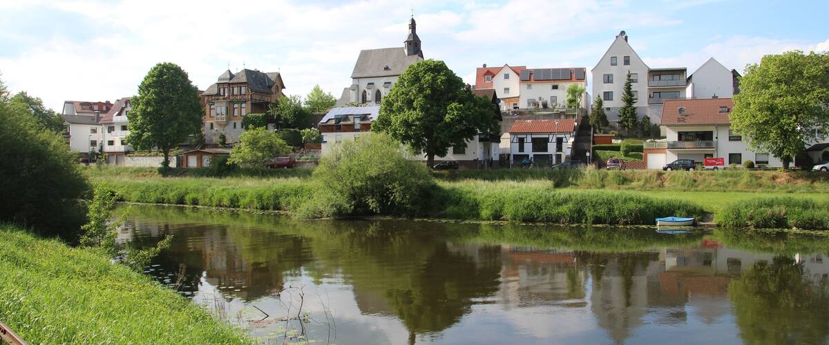 A calm river with a small boat, surrounded by lush greenery and traditional European buildings.