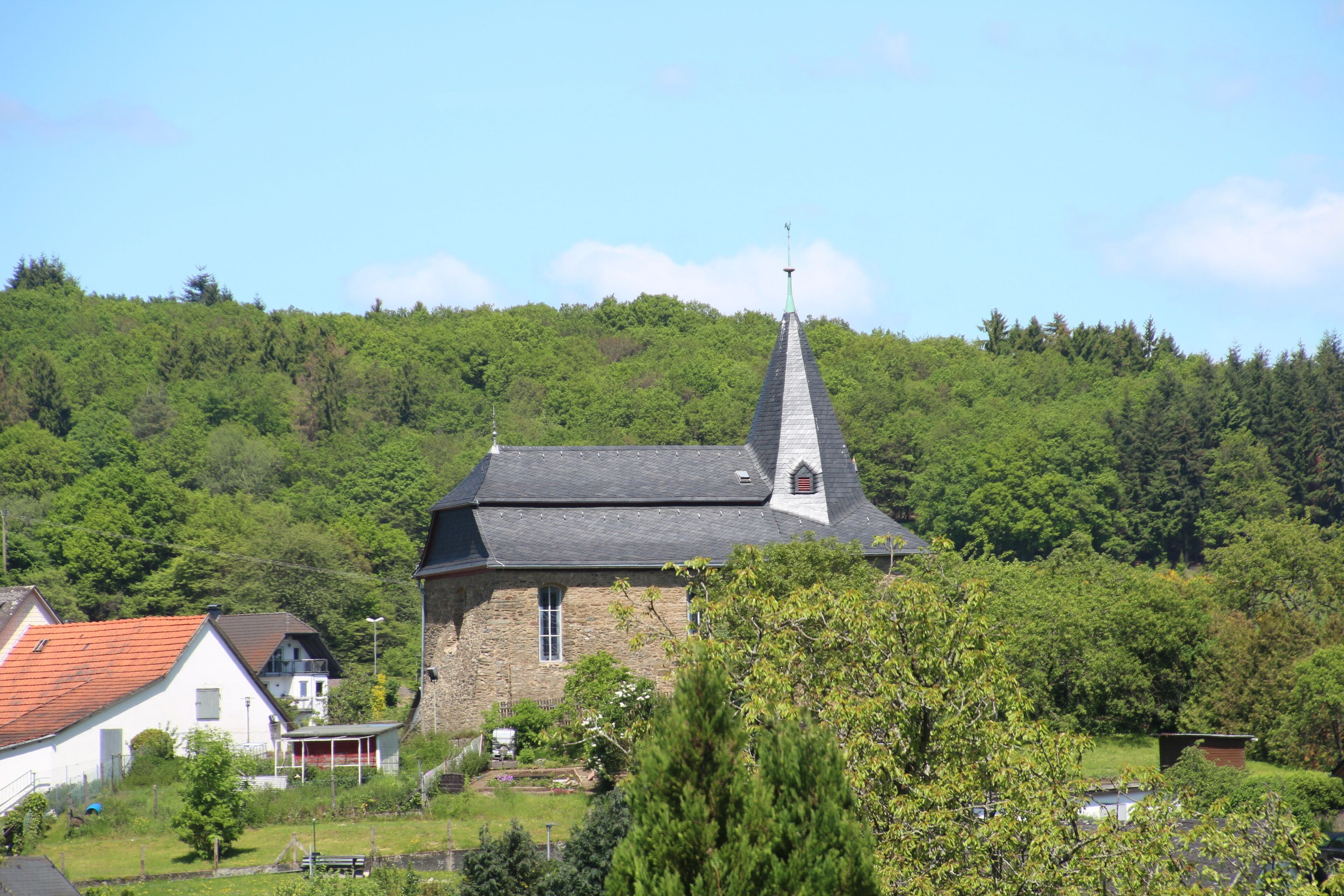 A church with a tall steeple, surrounded by lush greenery and a hill in the background.