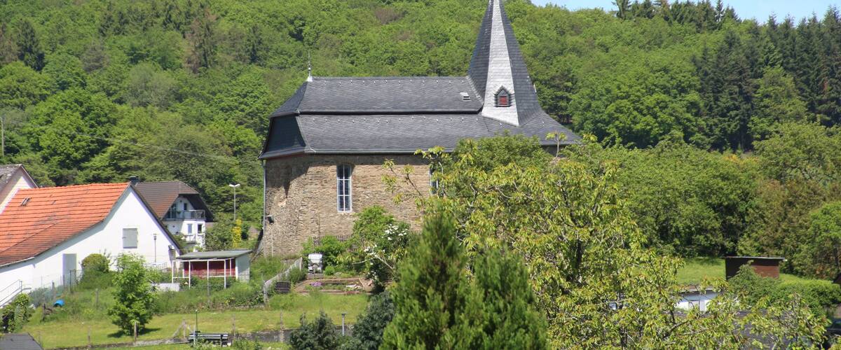 A church with a tall steeple, surrounded by lush greenery and a hill in the background.