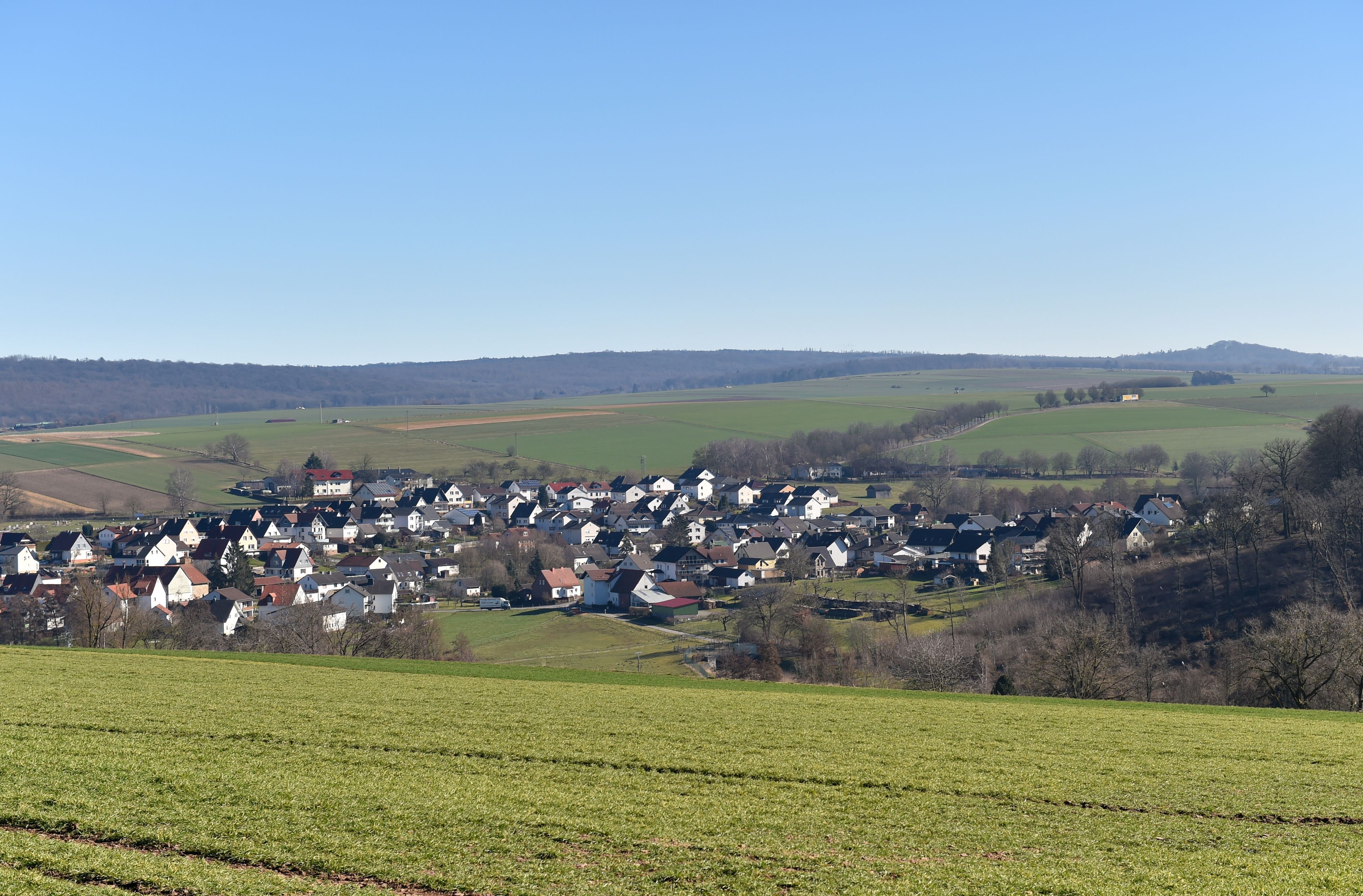Ein ländliches Dorf mit Häusern, die sich an einem Hügel schmiegen, umgeben von Feldern und weit entfernten Hügeln.