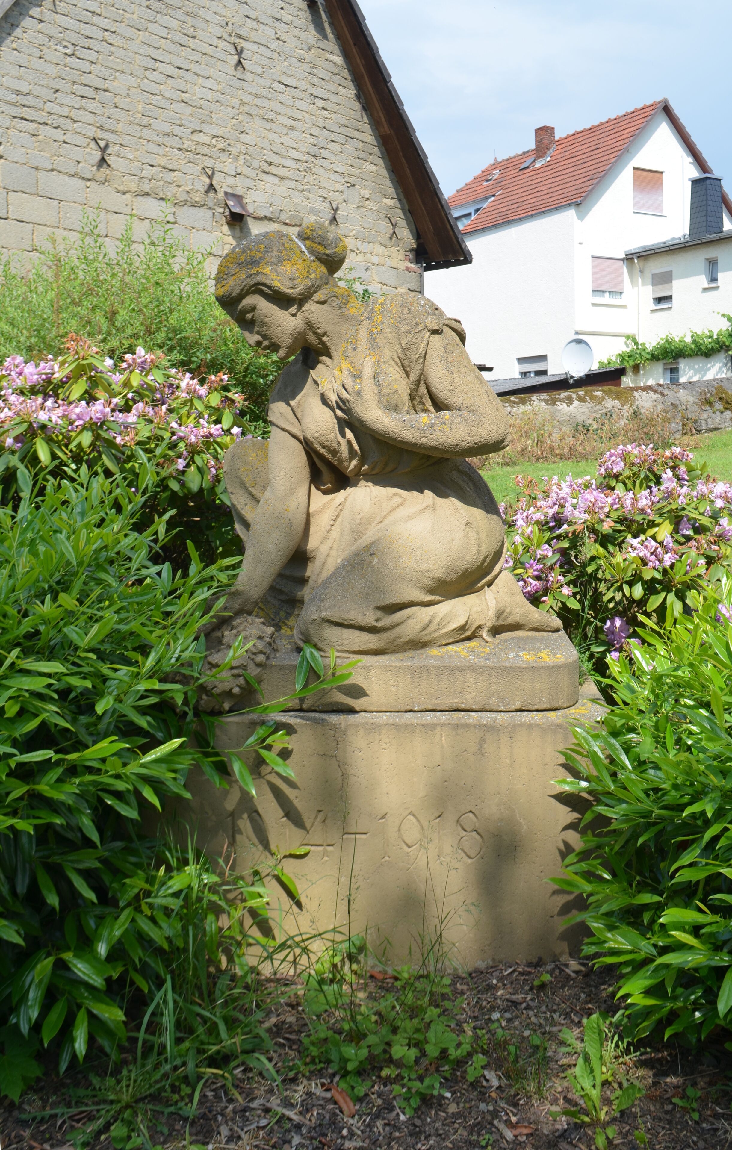 A stone sculpture of a figure in a garden with pink flowers and a building in the background.