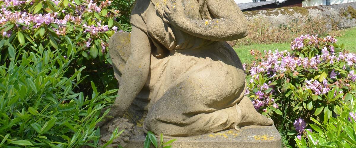 A stone sculpture of a figure in a garden with pink flowers and a building in the background.