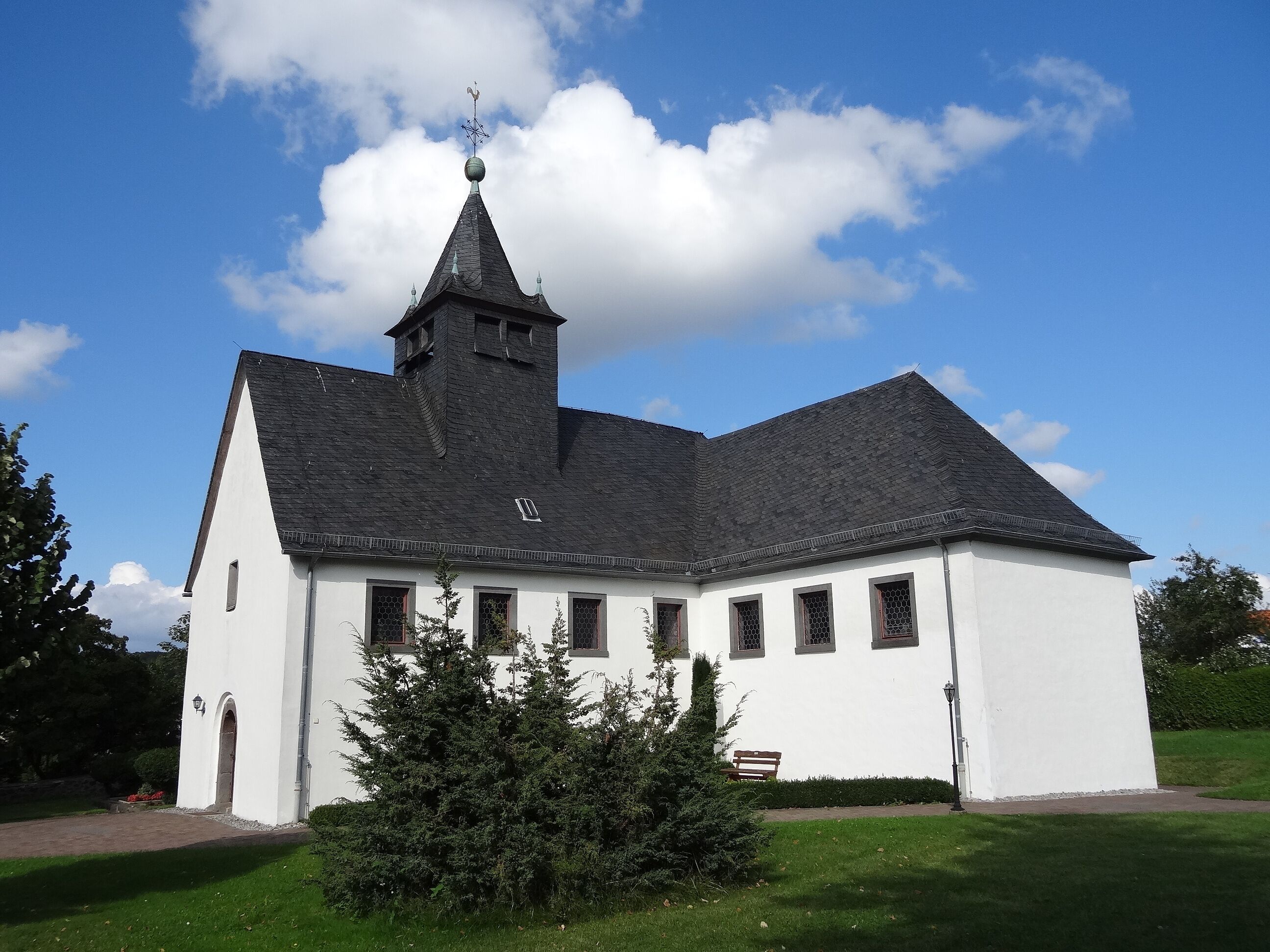 A white church with a black steeple and a weather vane, surrounded by green grass and trees.