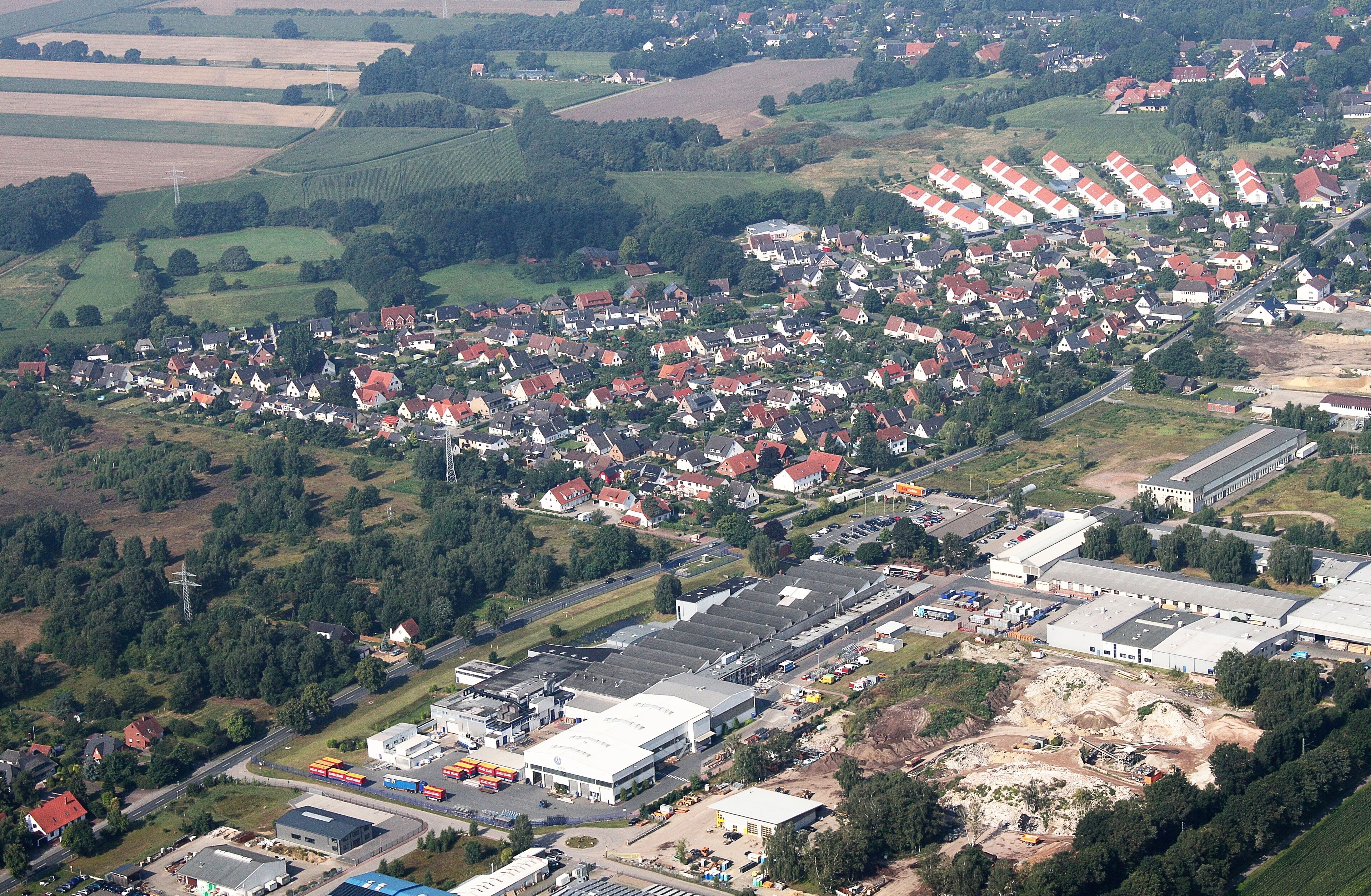An aerial view of a residential area with houses, a large industrial complex, and surrounding fields.