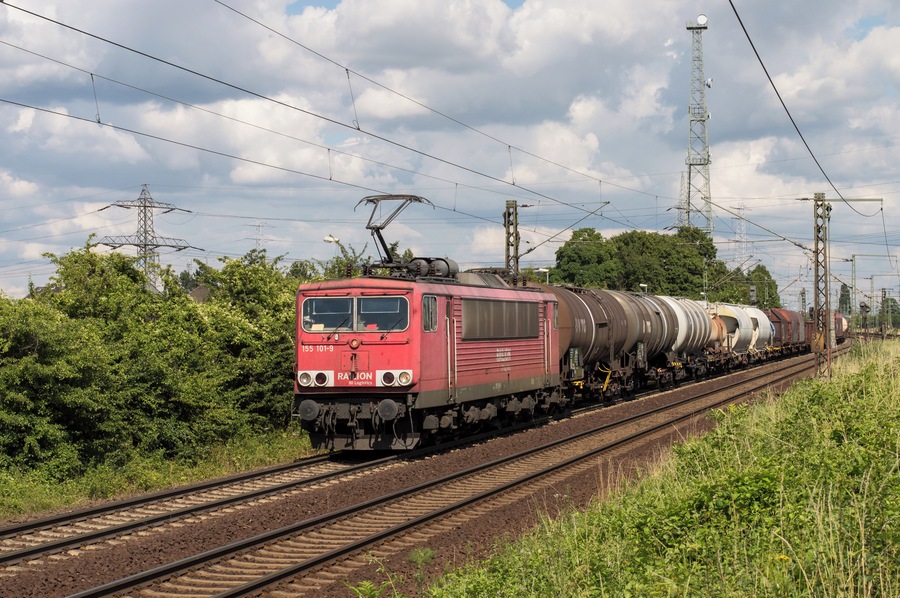 A red train with multiple tank cars traveling on tracks, surrounded by greenery and power lines.