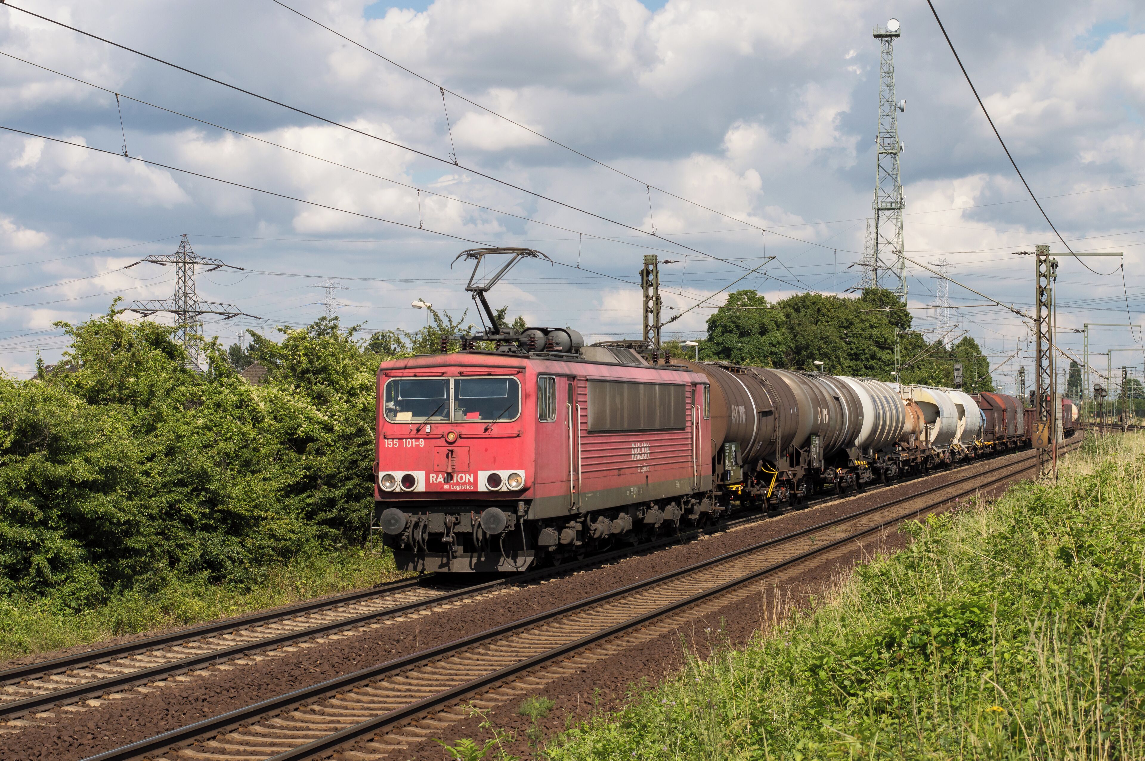 A red train with multiple tank cars traveling on tracks, surrounded by greenery and power lines.