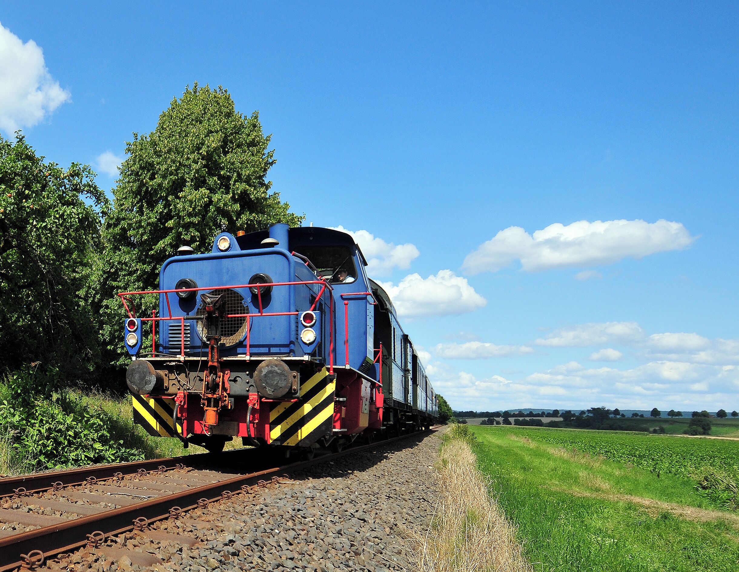 A blue train on tracks with a rural landscape and a clear sky.
