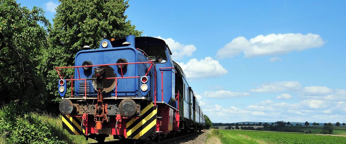 A blue train on tracks with a rural landscape and a clear sky.
