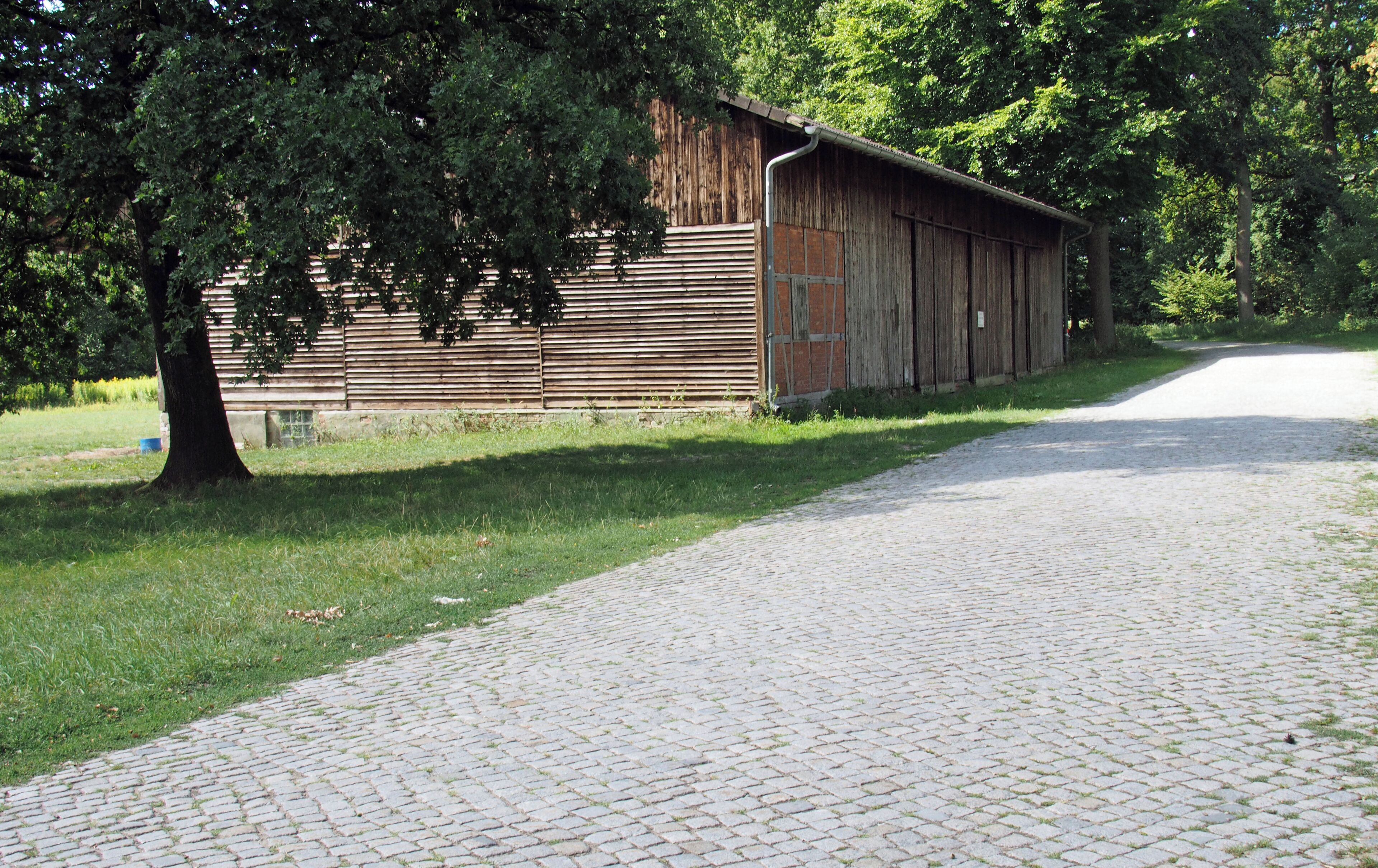 A rustic wooden barn with a cobblestone path leading to it, surrounded by lush greenery and trees.