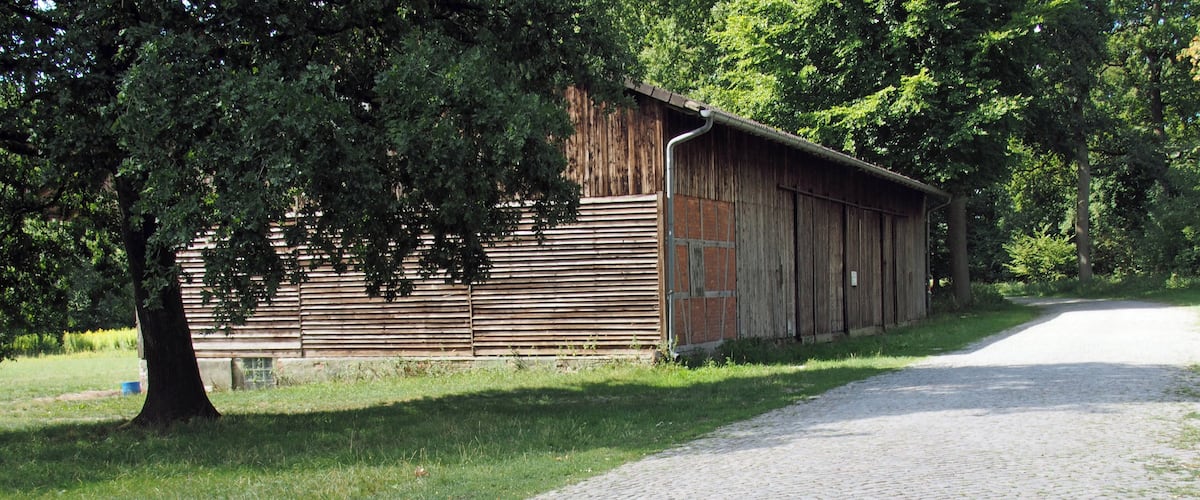 A rustic wooden barn with a cobblestone path leading to it, surrounded by lush greenery and trees.