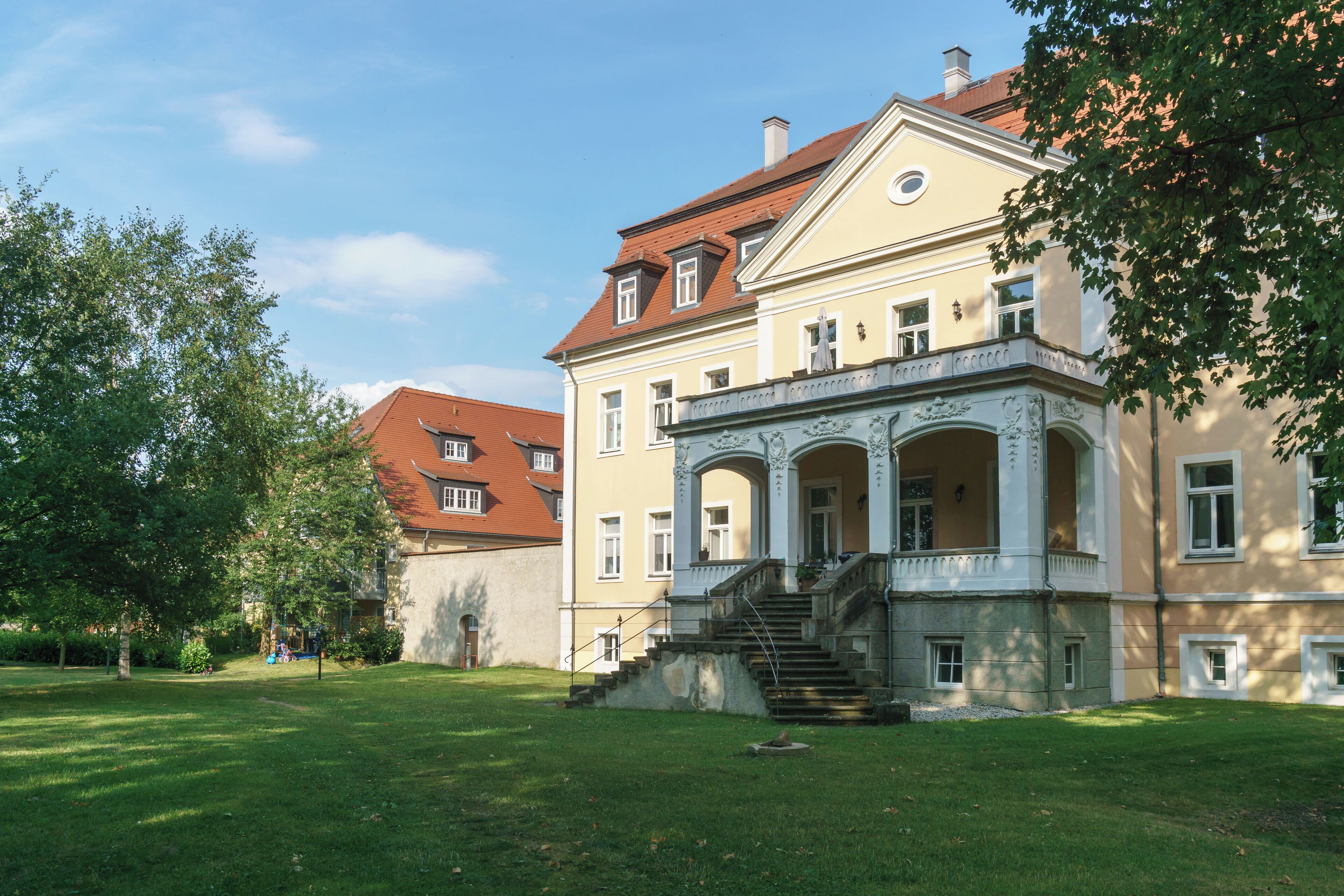 A historic building with a red roof, a large garden, and a staircase leading to the entrance.