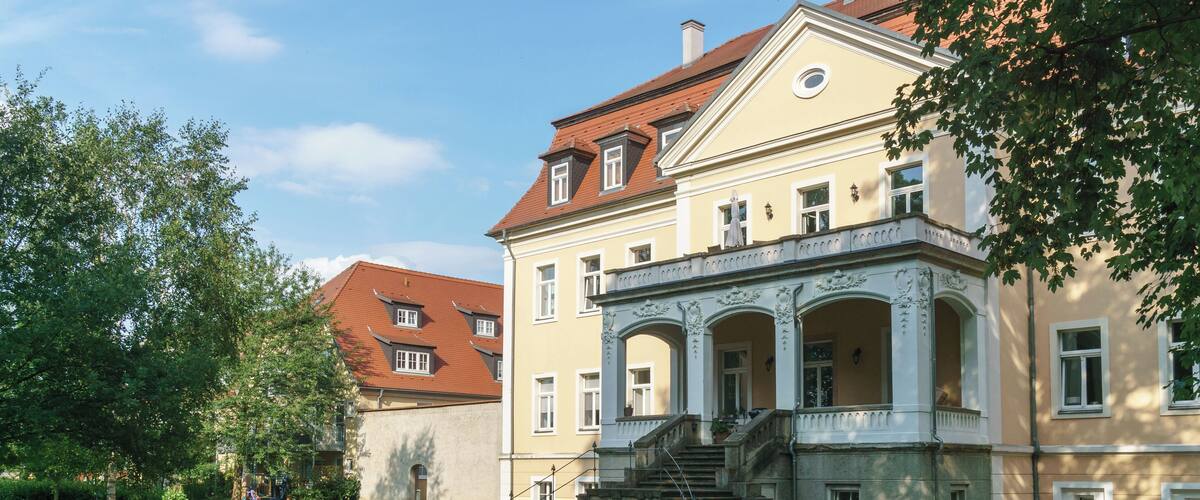 A historic building with a red roof, a large garden, and a staircase leading to the entrance.
