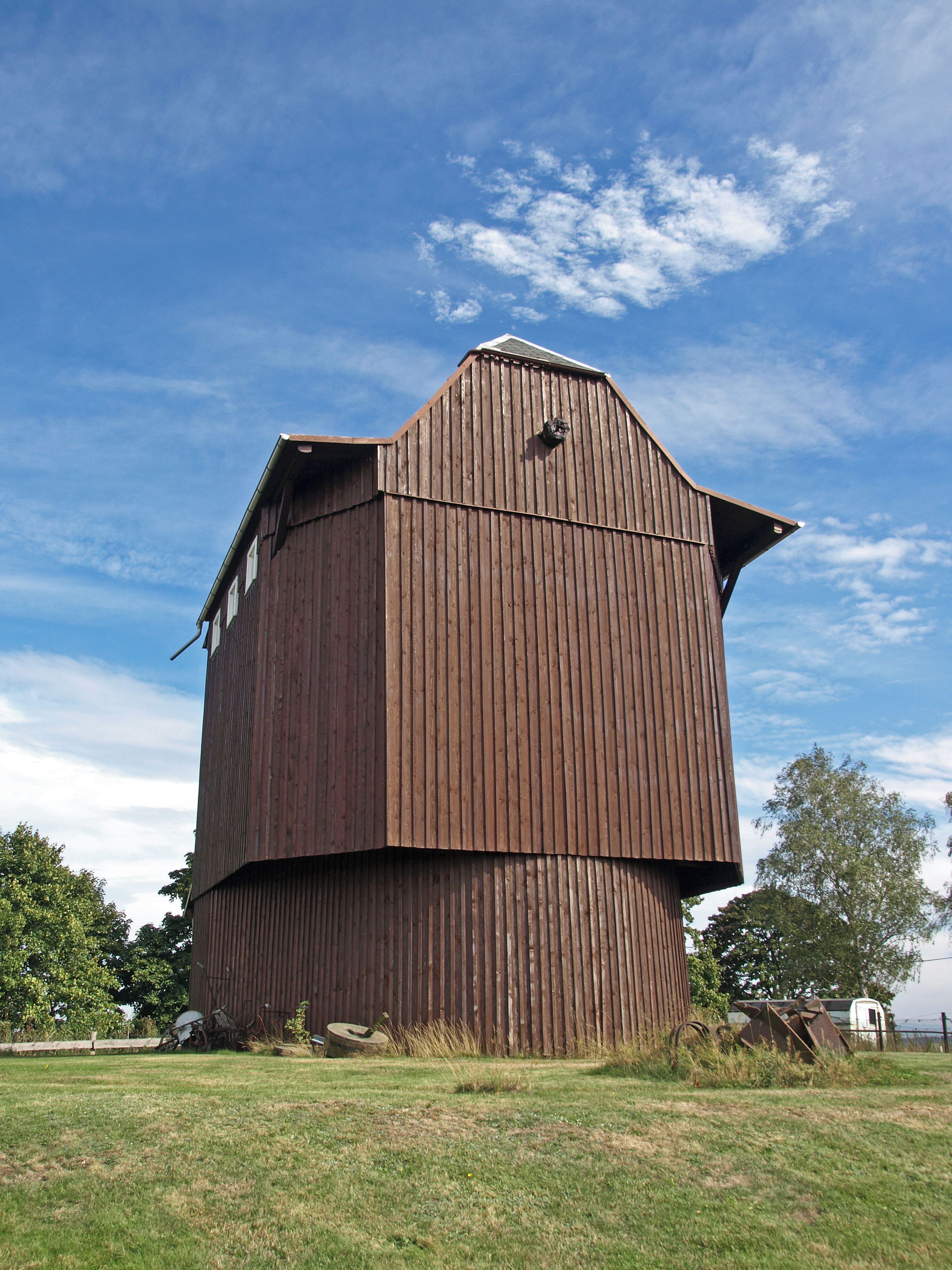 A large, wooden barn with a sloped roof, situated in a grassy field with trees in the background.