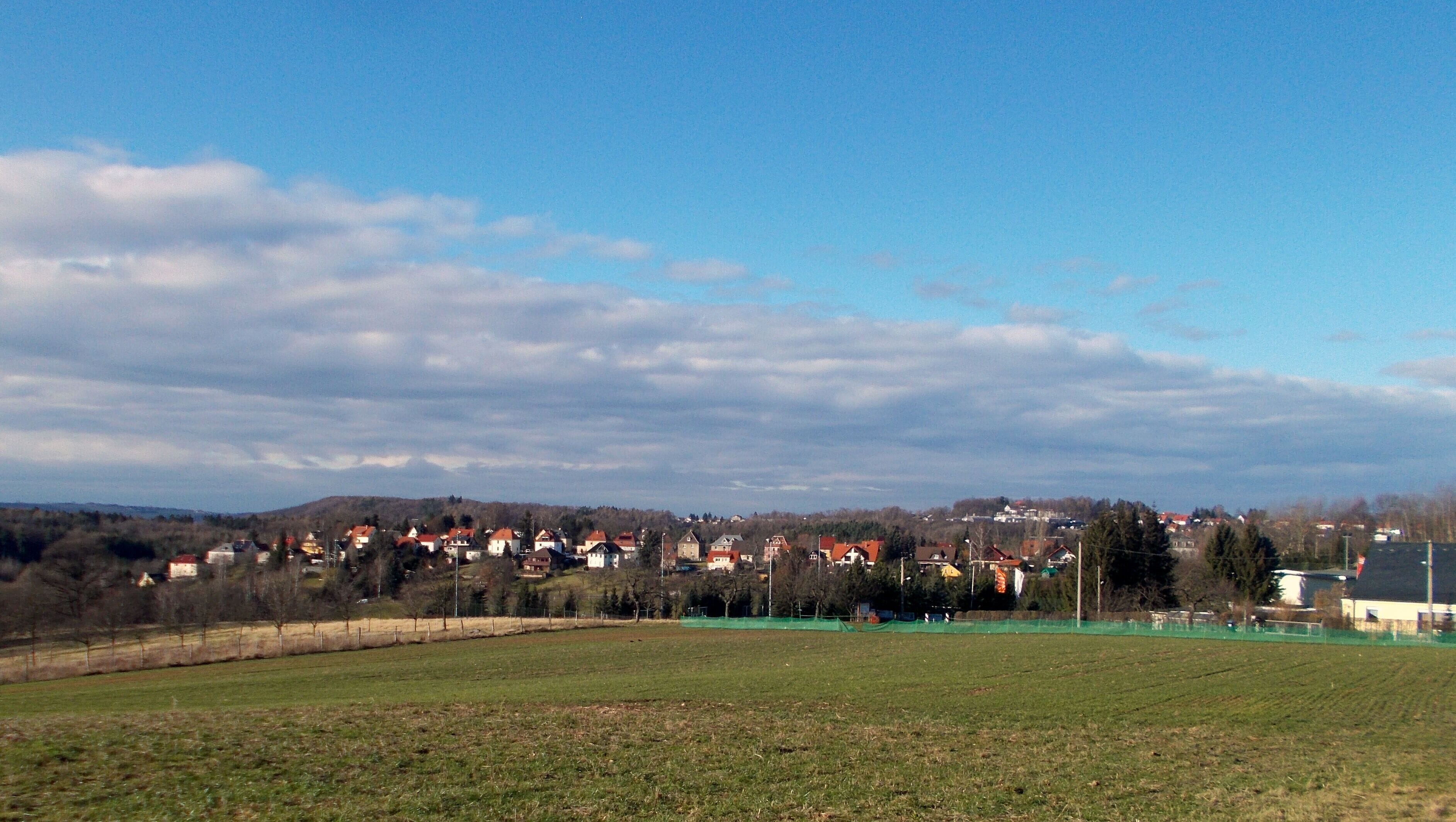 A rural landscape with a green field, a row of houses with red roofs, and a hill in the background.