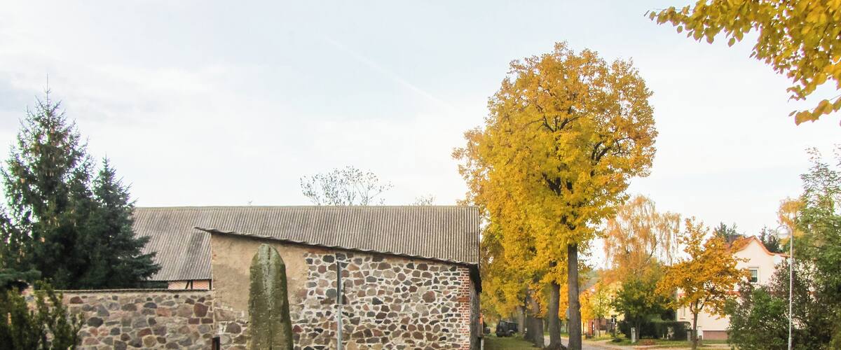 A stone building with a corrugated metal roof, surrounded by grass, fallen leaves, and autumn-colored trees.