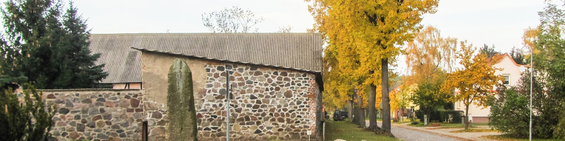 A stone building with a corrugated metal roof, surrounded by grass, fallen leaves, and autumn-colored trees.
