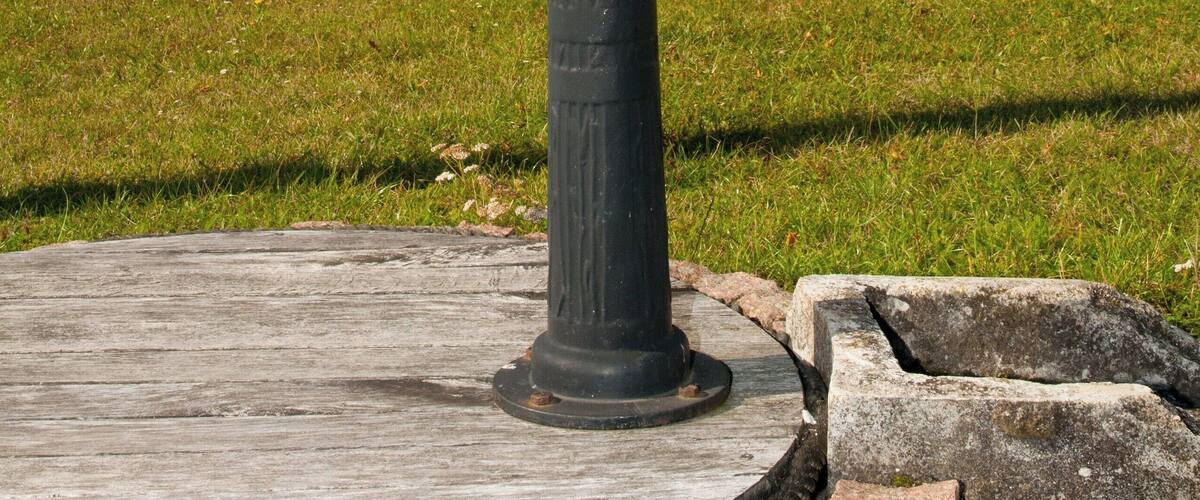 A metal fountain with intricate designs, surrounded by a circular stone base, set in a grassy area with trees displaying autumn foliage.