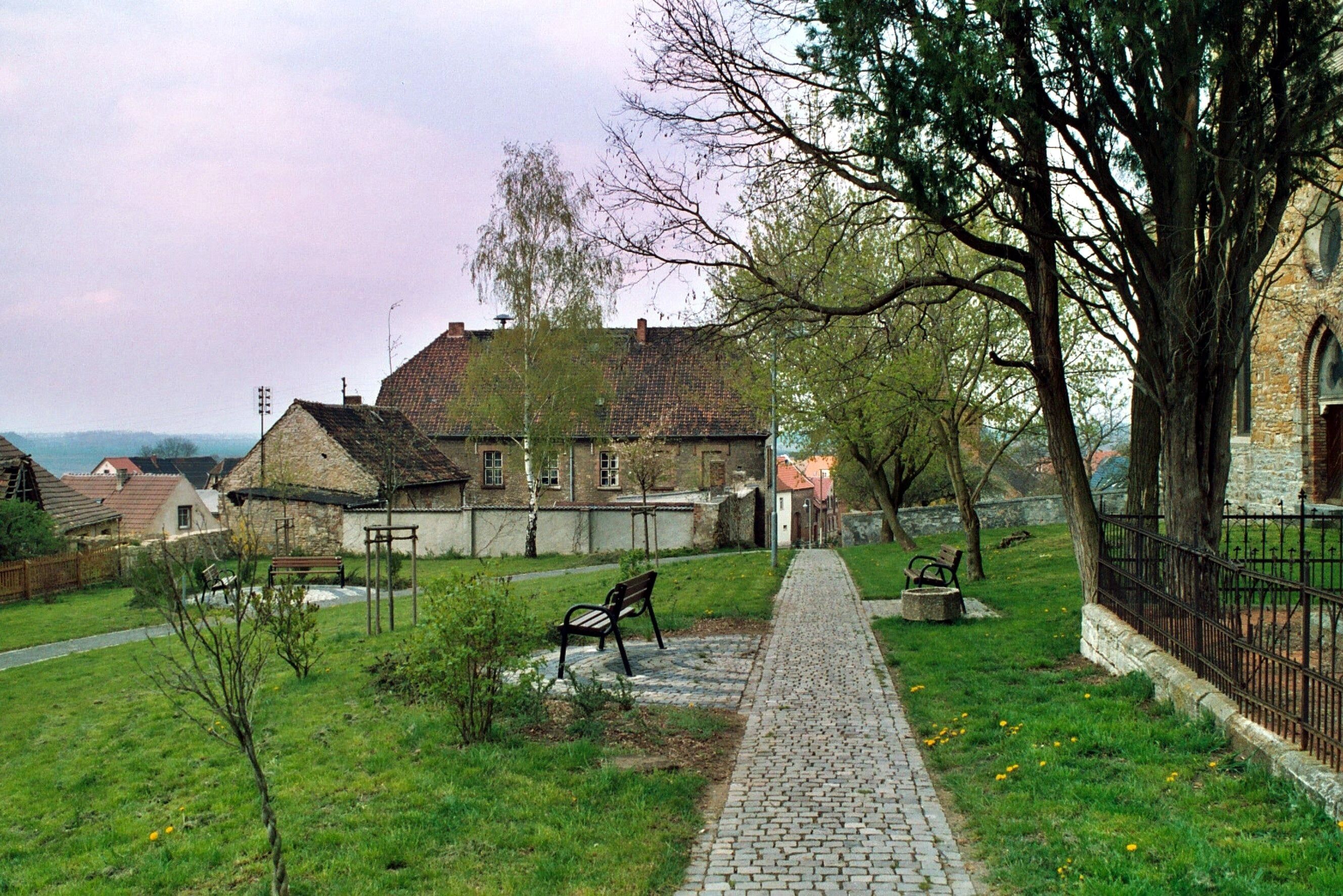A cobblestone path leading to a stone building with a red roof, surrounded by trees and a grassy area with benches.