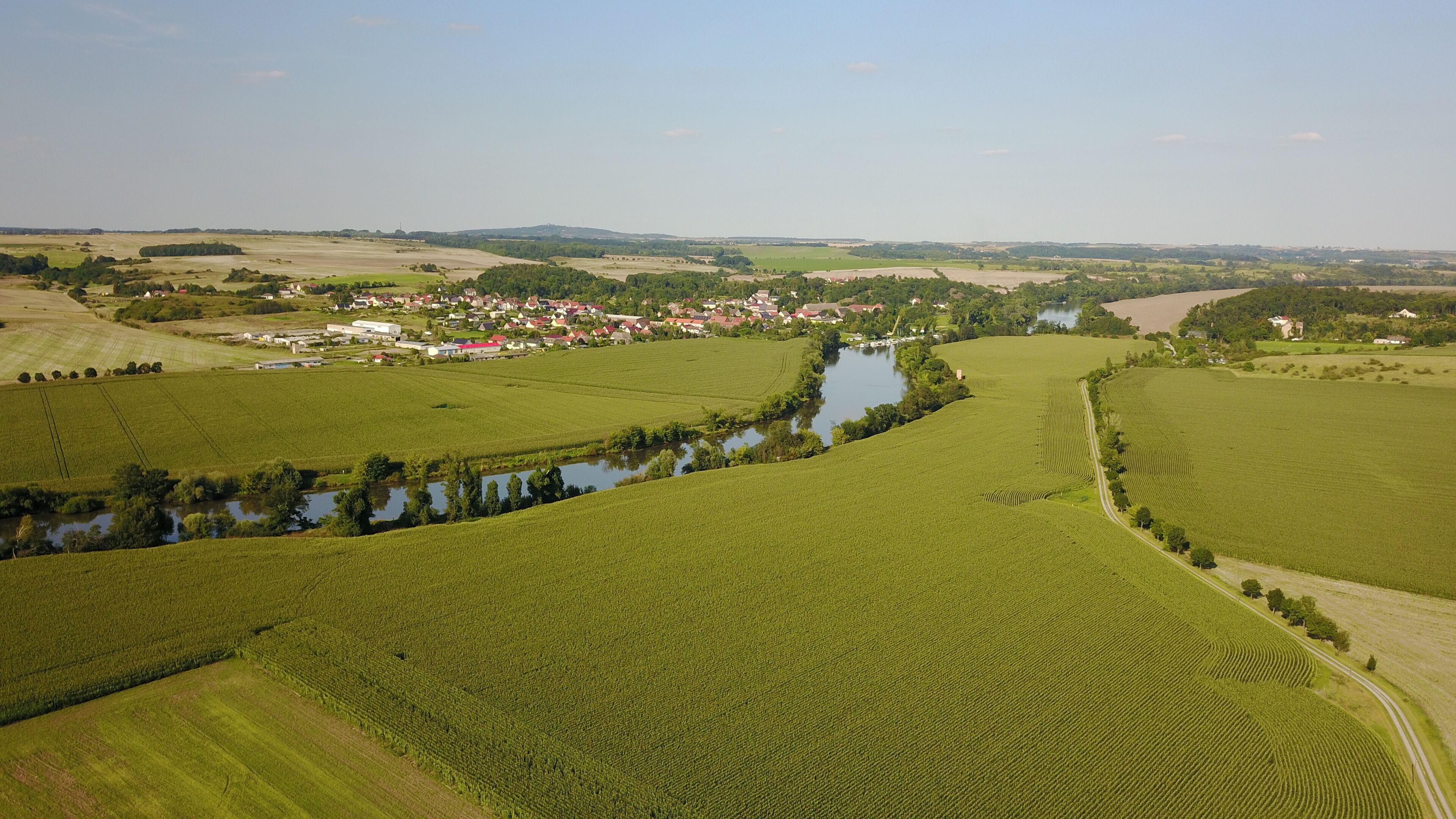 A river winding through green fields, a small village with red-roofed houses, and distant hills.