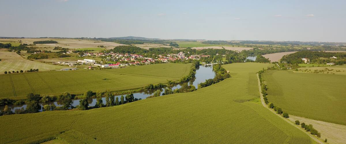 A river winding through green fields, a small village with red-roofed houses, and distant hills.