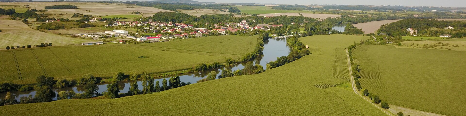 A river winding through green fields, a small village with red-roofed houses, and distant hills.