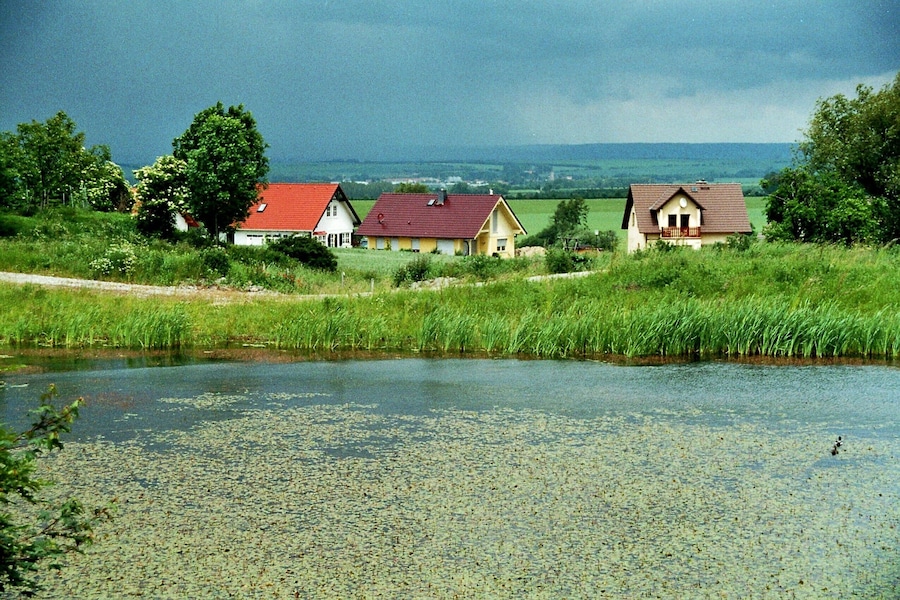 A rural landscape with a pond, three houses, and a cloudy sky.