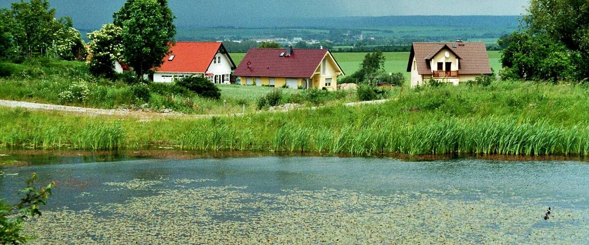 A rural landscape with a pond, three houses, and a cloudy sky.