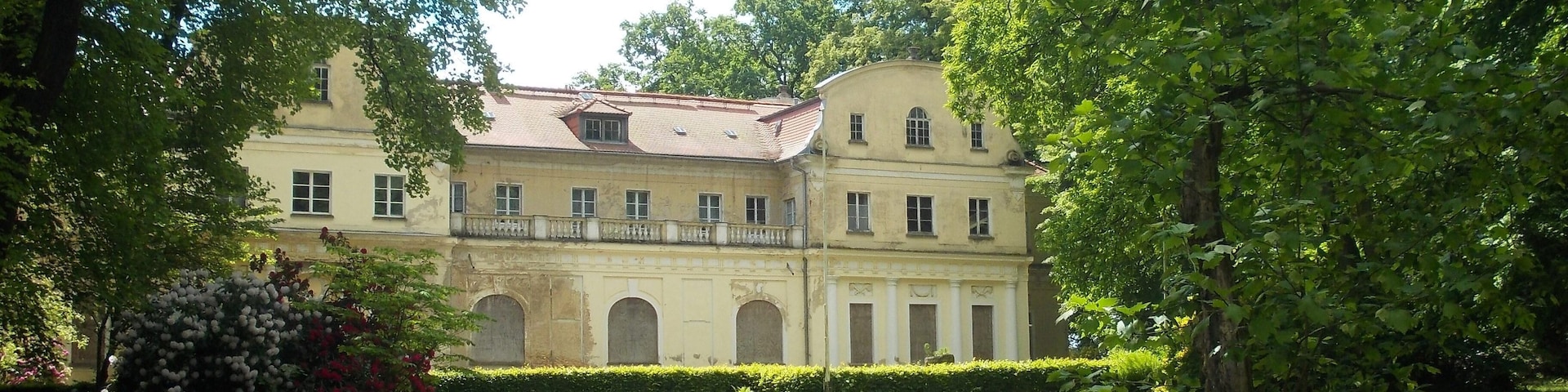 Un edificio grande y antiguo con ventanas arqueadas y una fachada simétrica, rodeado de abundante vegetación y un césped cuidado.
