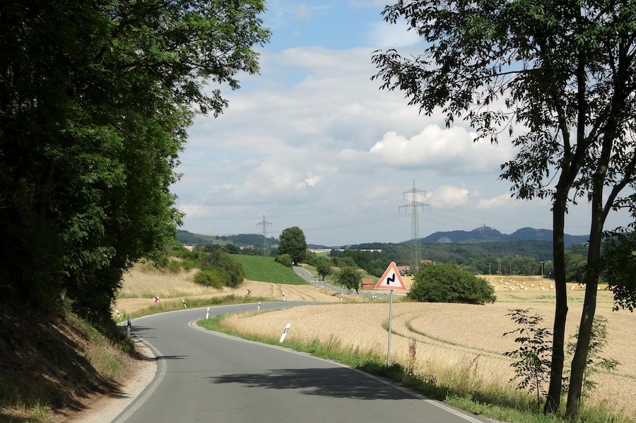 Uma estrada rural sinuosa com uma curva acentuada, rodeada por campos e árvores.