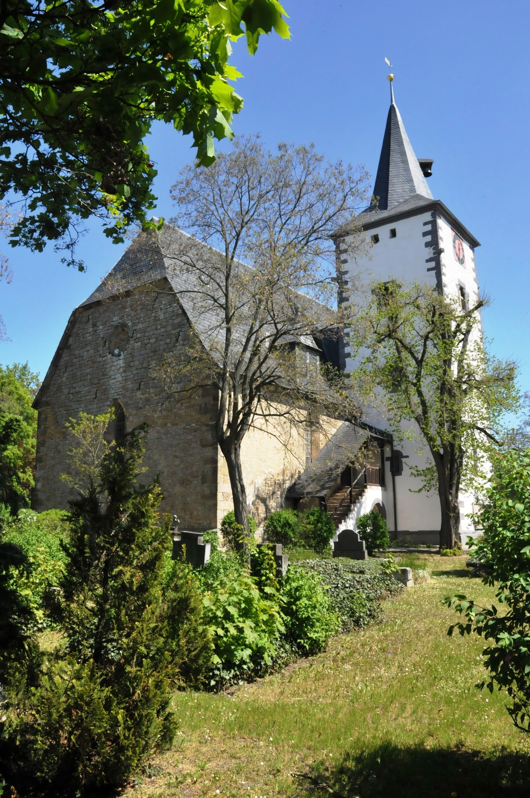 A historic church with a tall steeple and a clock tower, surrounded by lush greenery and trees.