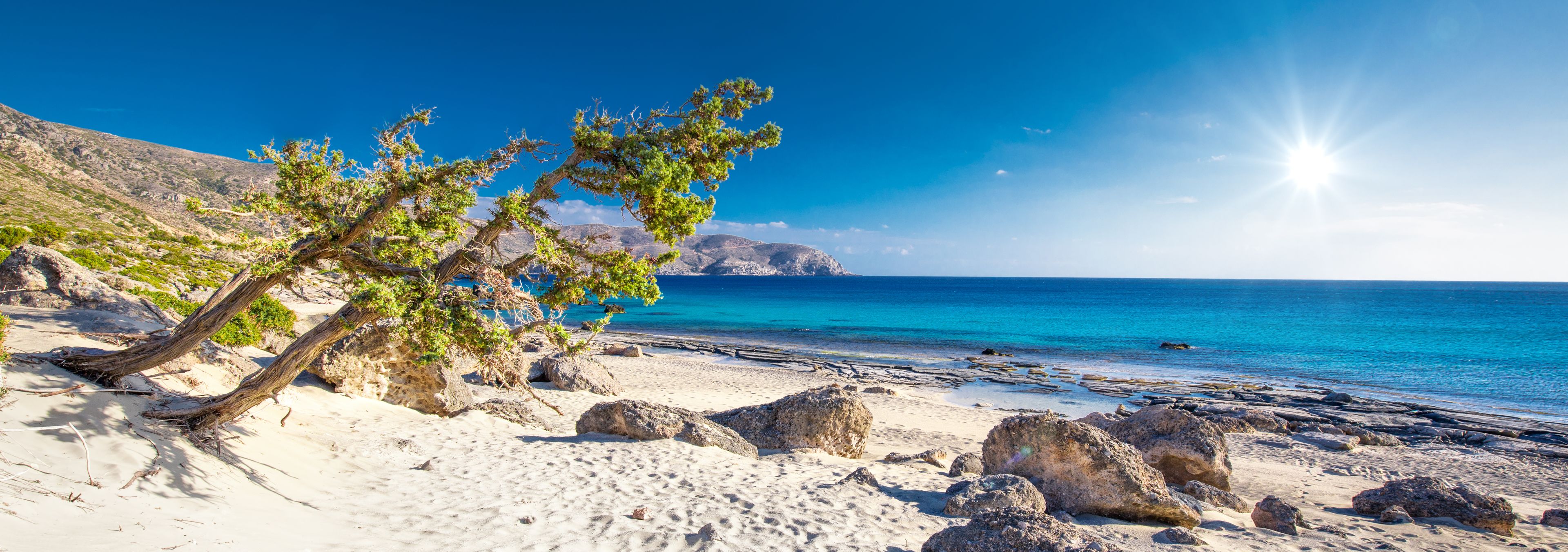 A beach with white sand, scattered rocks, and a clear blue sea under a bright sun.