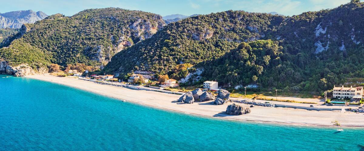 A coastal landscape with a sandy beach, clear turquoise waters, and a backdrop of green hills.