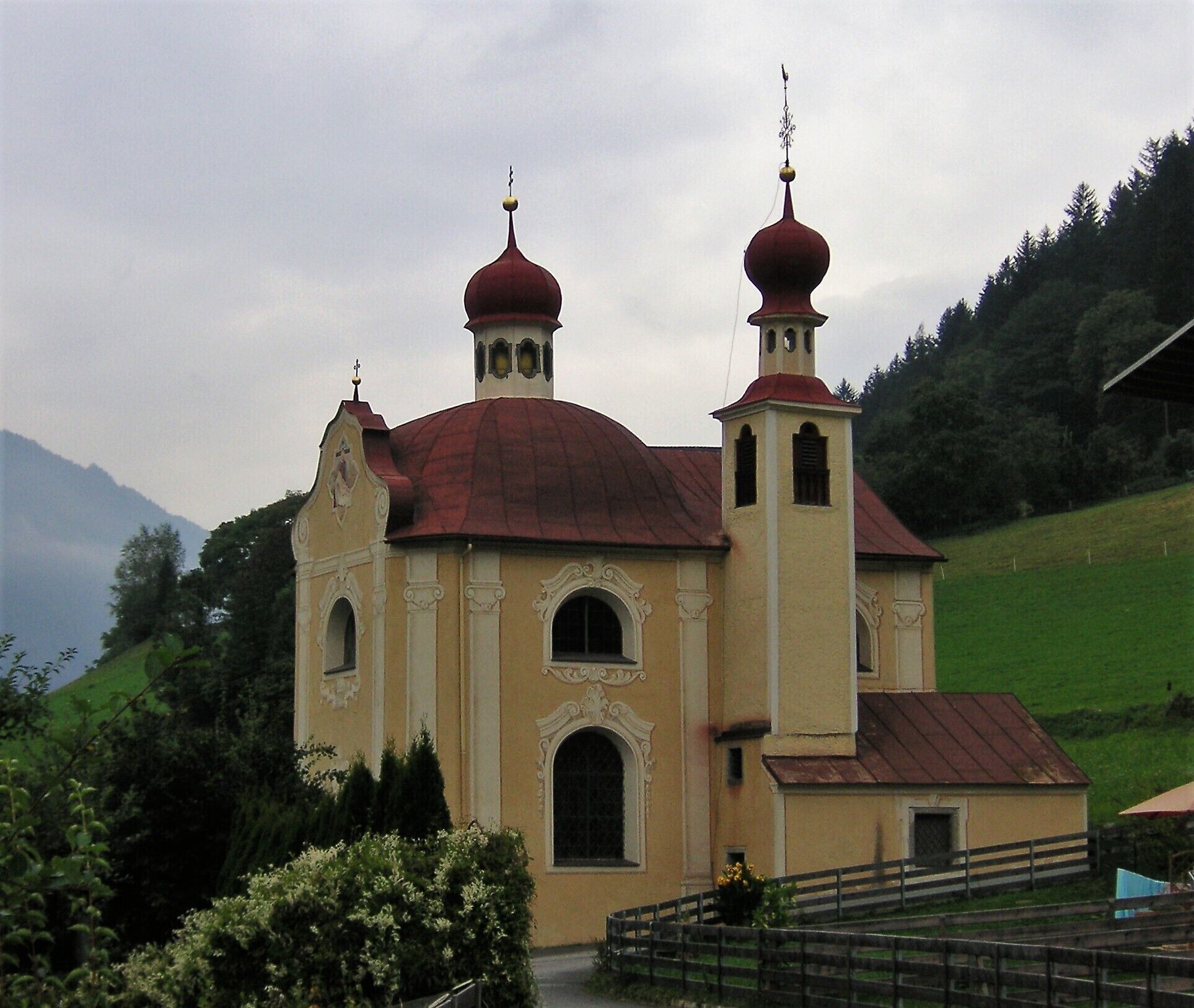 A church with a red dome and two bell towers, surrounded by greenery and hills.