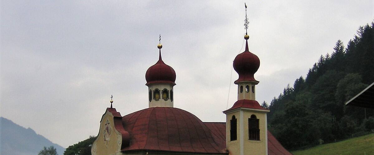 A church with a red dome and two bell towers, surrounded by greenery and hills.